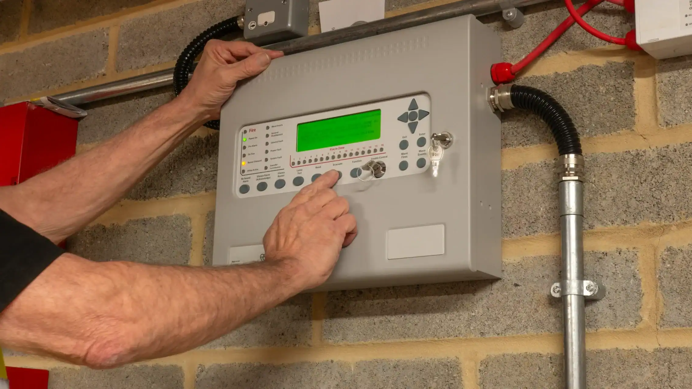 A person operates a wall-mounted control panel with a green display and multiple buttons in a room with exposed brick walls and wiring.