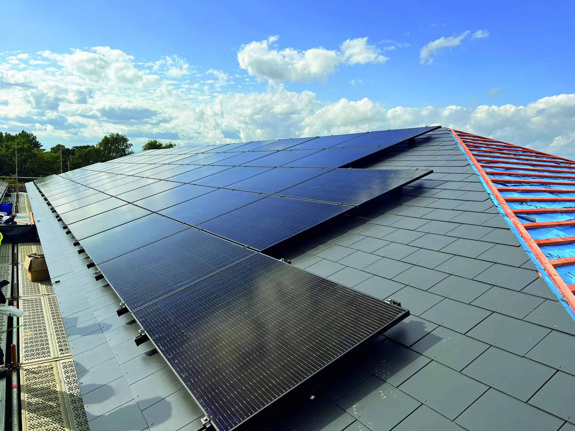 Rows of solar panels installed on a sloped rooftop under a partly cloudy sky, with construction materials visible along the roof edge.