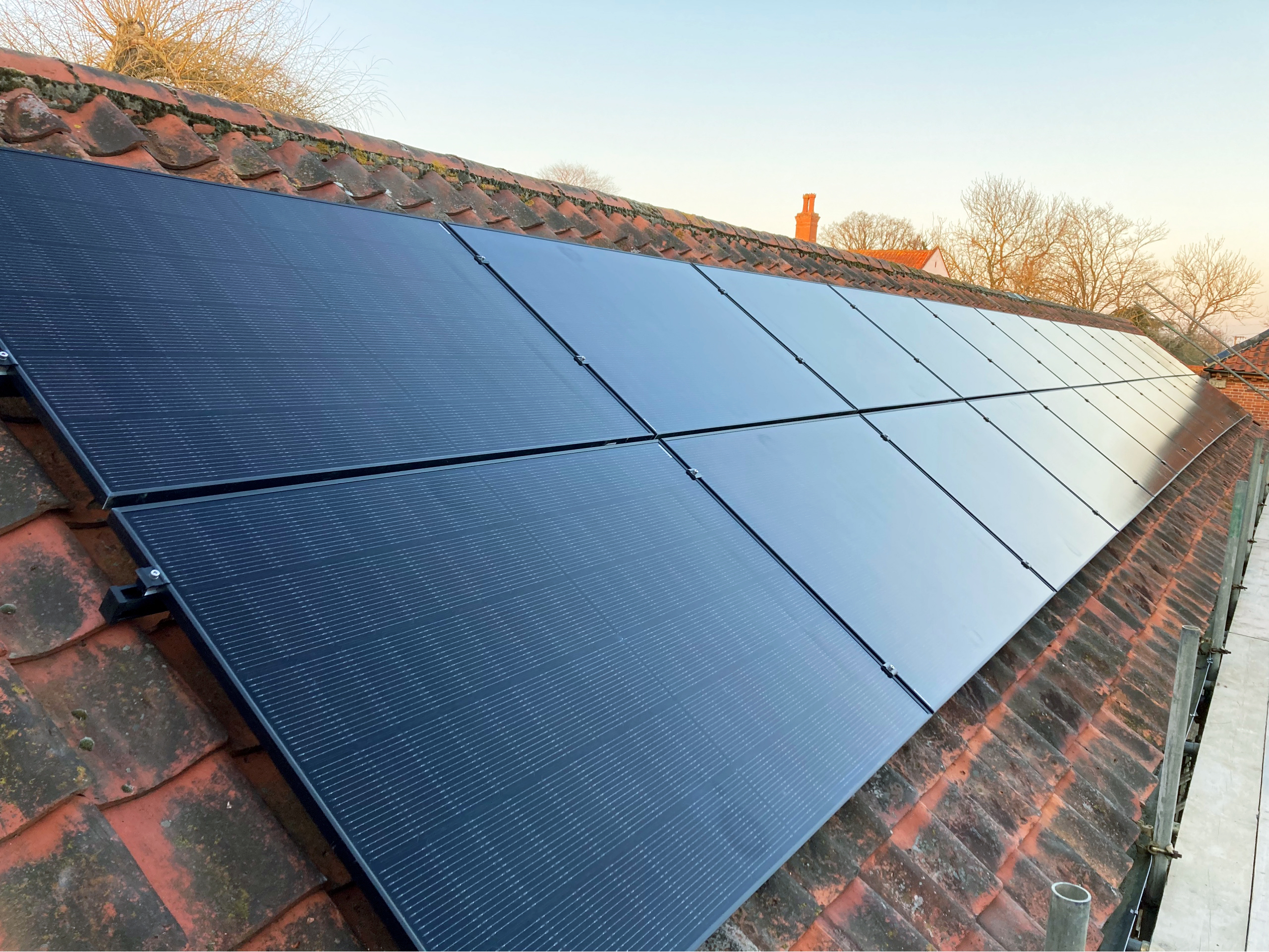 Solar panels fitted on a sloping, red-tiled roof with trees and a clear sky in the background.
