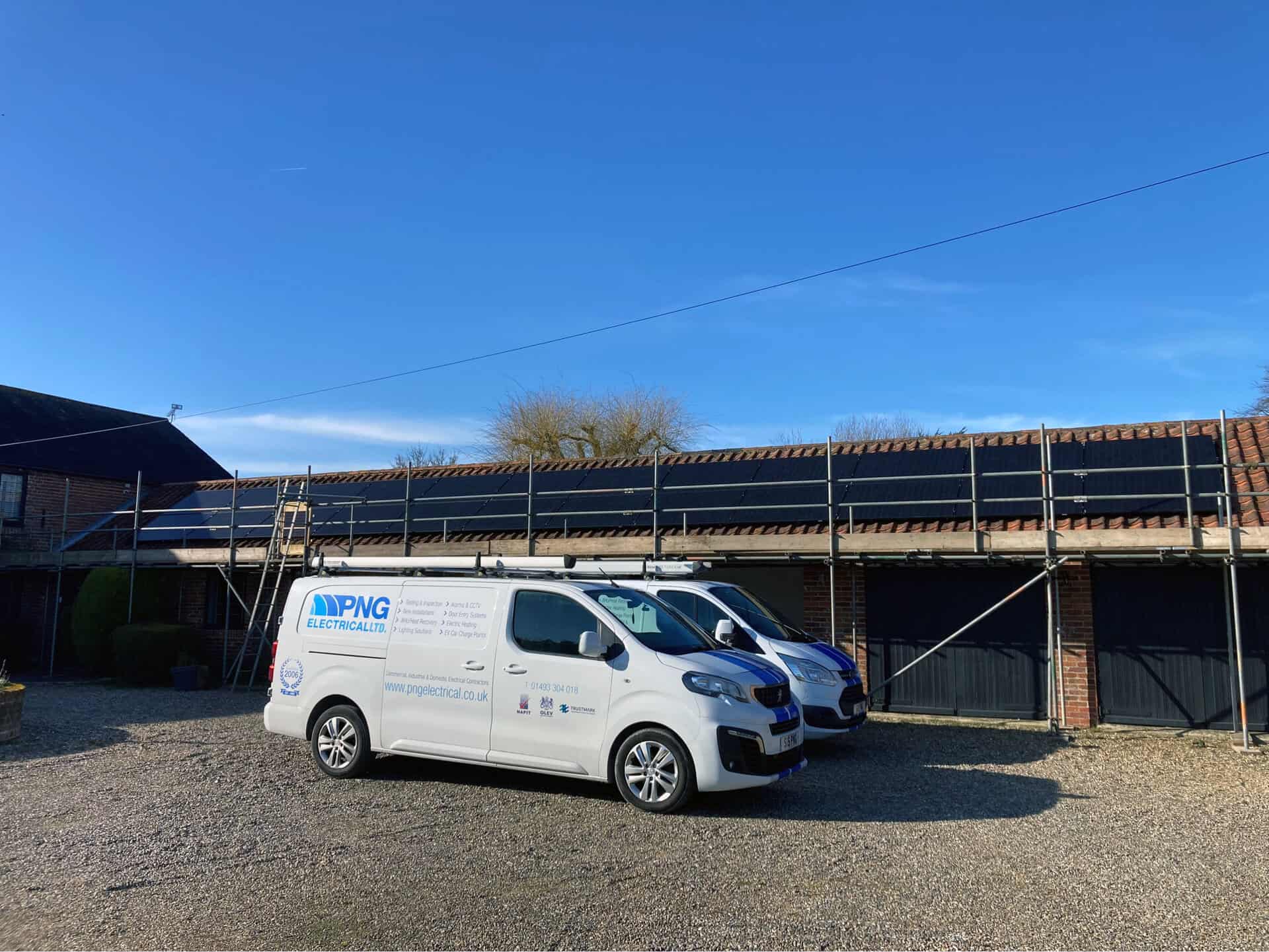 Two white vans are parked in front of a building with scaffolding and solar panels installed on its roof under a clear blue sky.