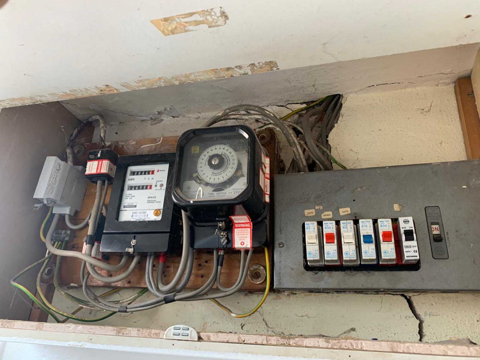 Electrical control panel with wiring, analogue timer, circuit breakers, and switches mounted on a wall inside a utility cupboard.