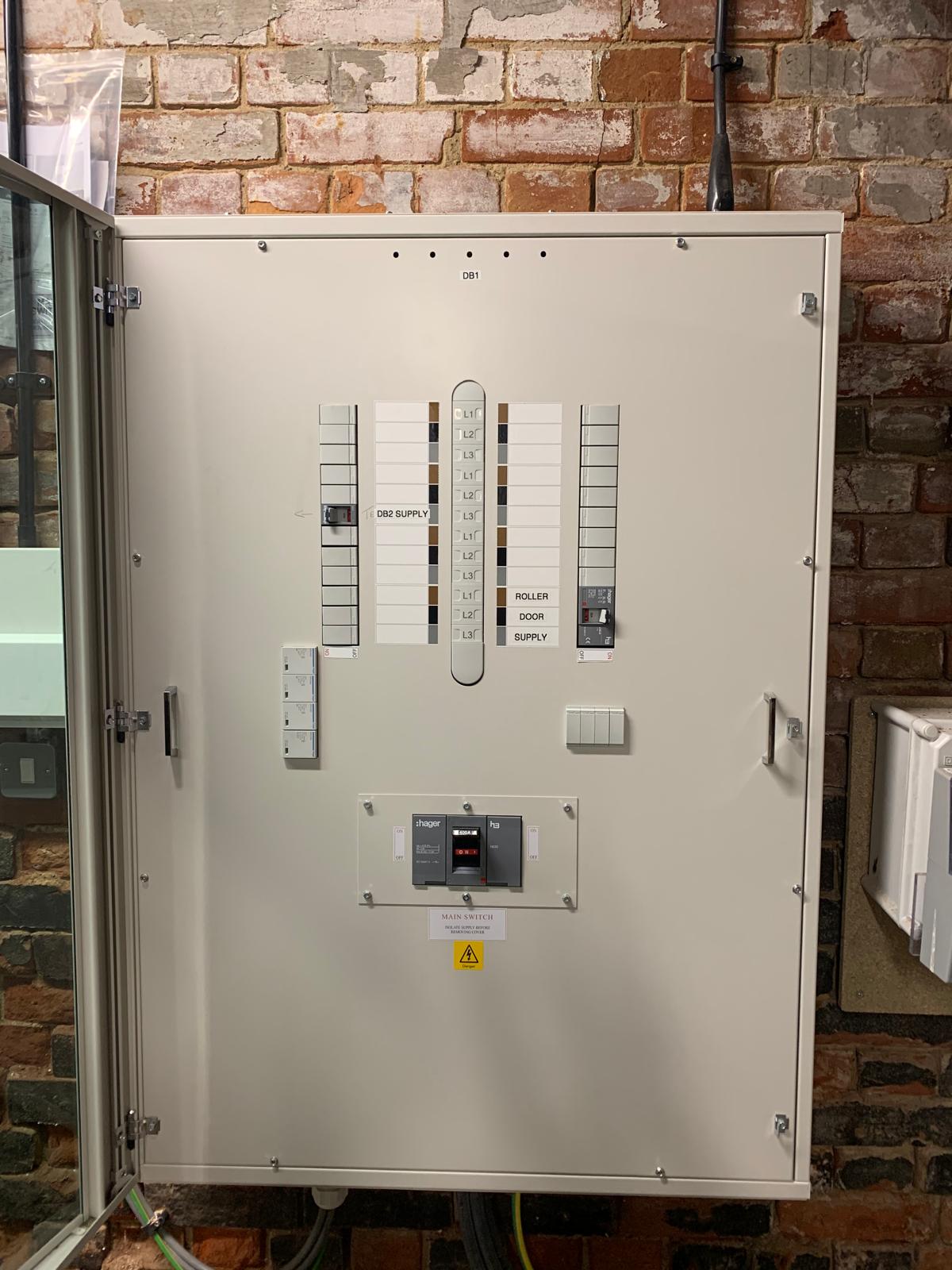 An electrical distribution board with labelled circuit breakers is mounted on a brick wall with its door open.