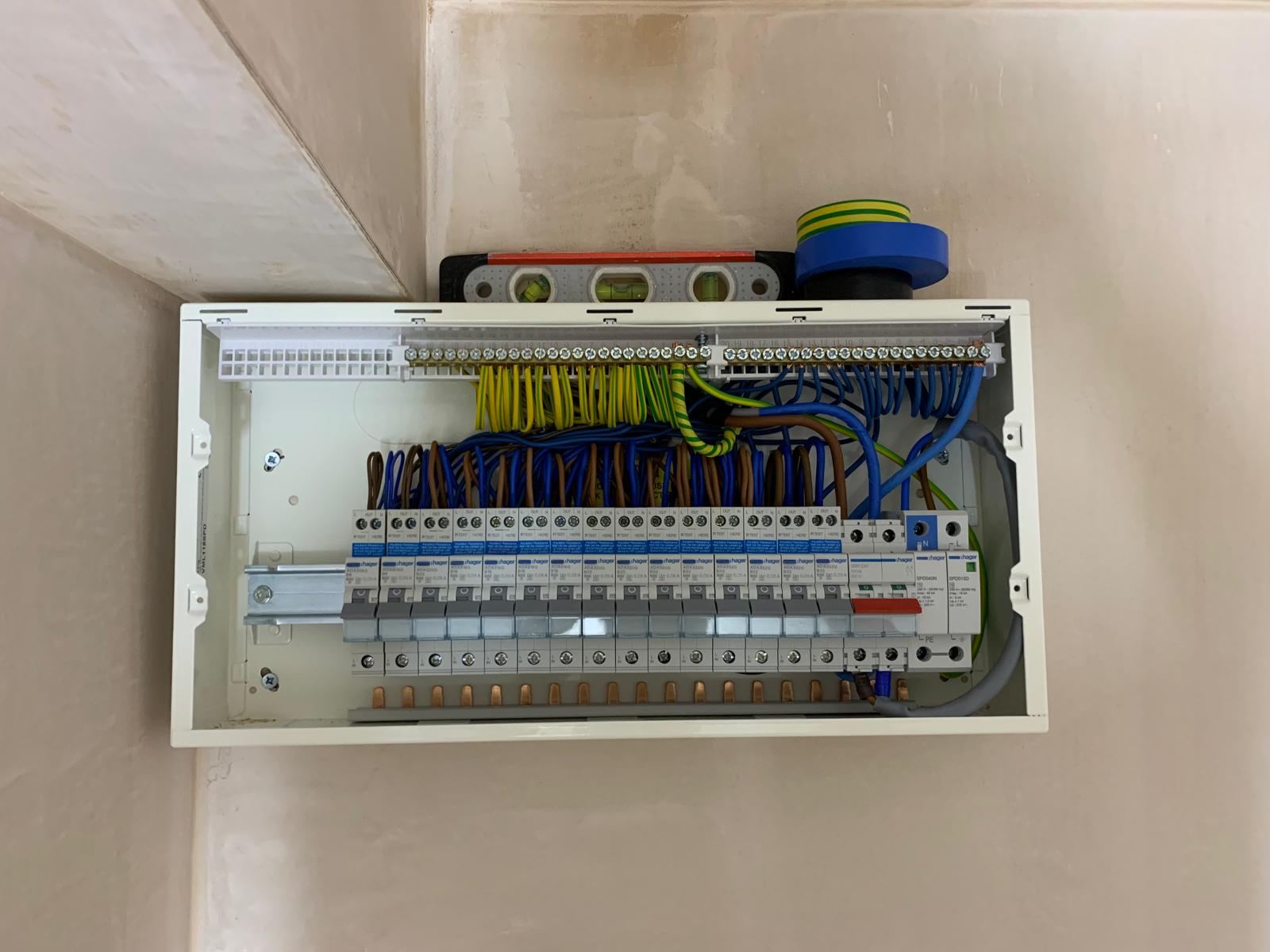 An open electrical distribution board shows circuit breakers, organised wiring, and a spirit level with tape on top.