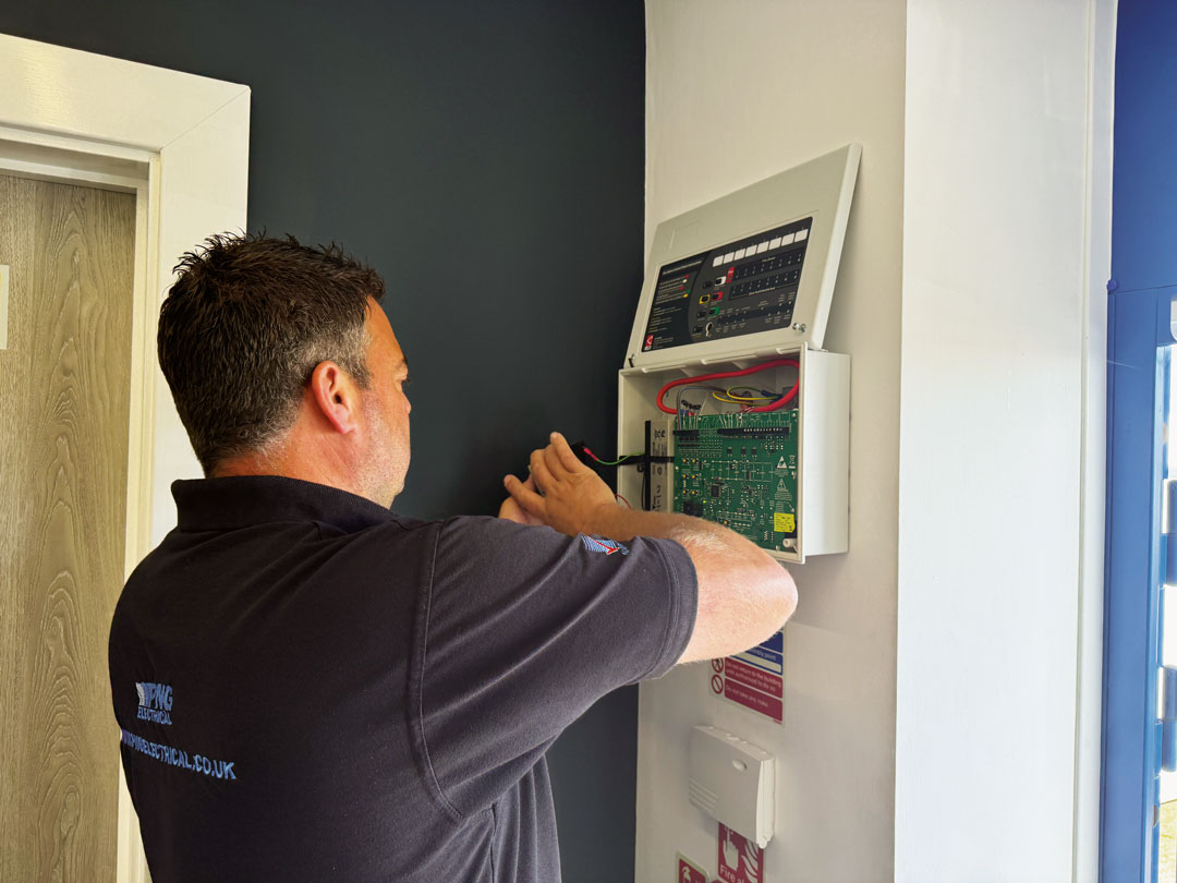 A person in a black polo shirt uses a screwdriver to work on an open electrical control panel mounted on a white wall.