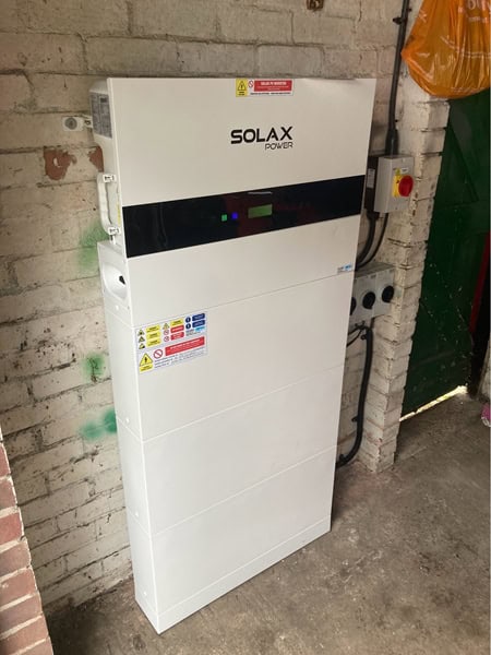 A white Solax Power battery storage unit is mounted on a brick wall in a utility area, with warning labels and a control switch nearby.